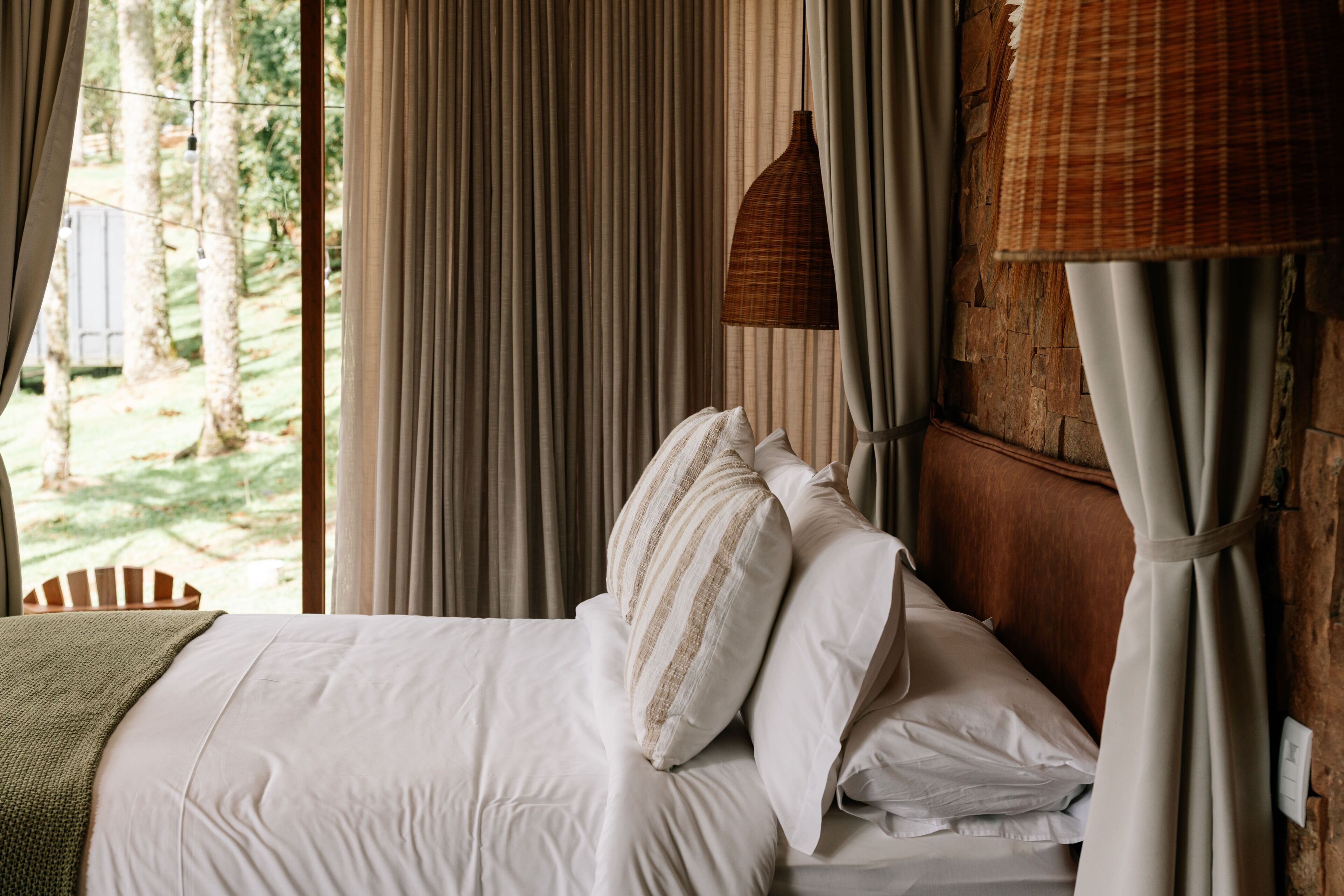 Uma cama com roupa de cama branca e almofadas listradas, cortinas e um candeeiro suspenso num quarto com vista para árvores.