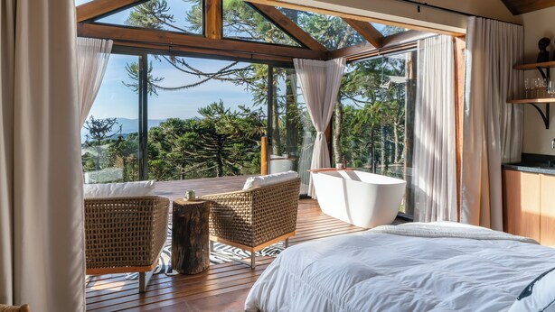 A bedroom with a large bed, a wooden deck, a white bathtub, and a view of trees and mountains. - Bulawayo Lodge (Urubici)