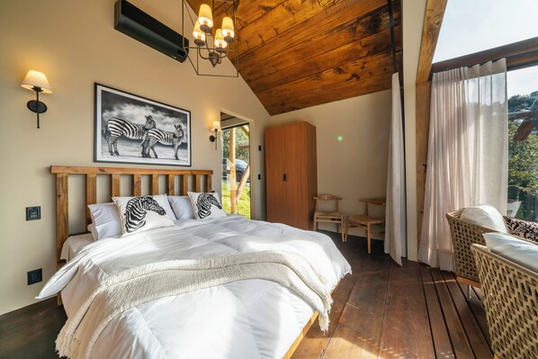 A bedroom with a large bed, zebra-themed pillows, a wooden headboard, a wooden ceiling, a wall-mounted lamp, and a view of greenery outside. - Bulawayo Lodge (Urubici)