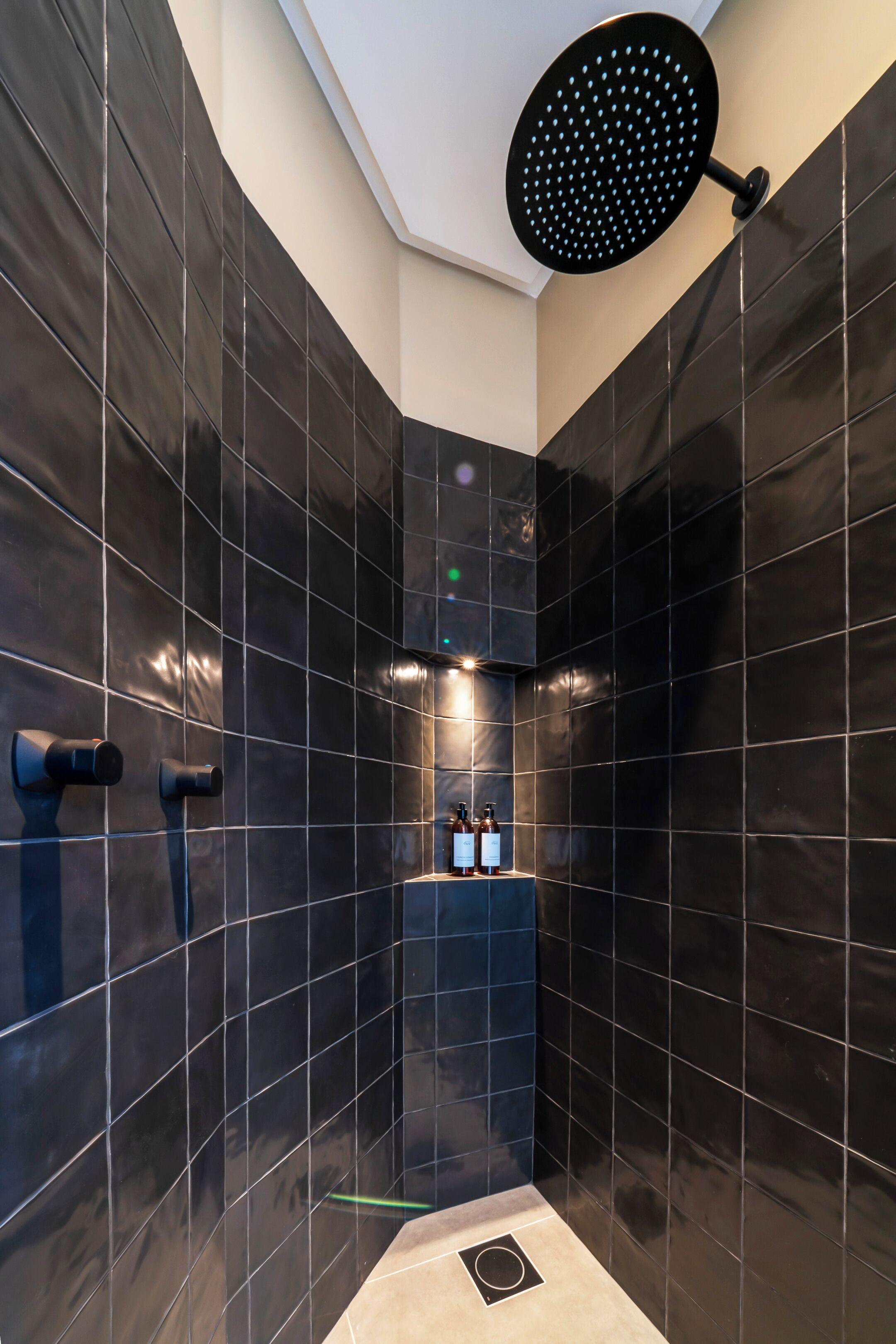 A modern shower with a circular head, black tiles, and a small shelf with bottles.