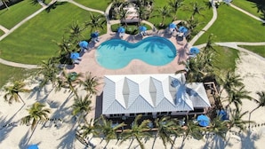 Eine Vogelperspektive zeigt ein Resort mit einem zentralen Schwimmbecken, umgeben von Palmen und einem Gebäude mit einem geometrischen Dach.