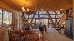 A cozy cabin interior with wooden walls and a large window, a dining area with a Christmas tree, and a staircase leading to an upper level.