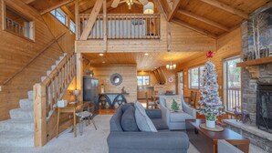 A spacious log cabin interior with a staircase leading to a loft, a cozy living area with a fireplace, and a decorated Christmas tree.