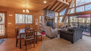 A cozy cabin interior with a dining table, chairs, a fireplace, a sofa, and large windows offering a view of the surrounding forest.