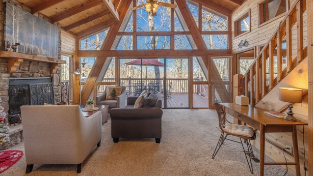 A spacious cabin interior with a high ceiling, wooden beams, a fireplace, a dining area, and large windows offering views of the outdoors.