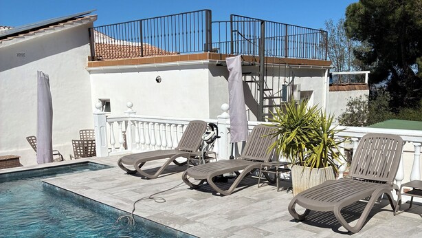 A swimming pool with clear water, surrounded by lounge chairs and umbrellas, with a white building and a tree in the background. - Villa with heated pool and private padel court (Pinos de Alhaurín)