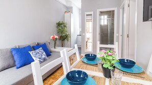 A modern living room with a grey sofa, blue cushions, a dining table with blue plates and bowls, and a potted plant with pink flowers. - Cozy apartment close to Atocha and Retiro, Madrid - Cuore Apartments (Madrid)