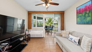 Una sala de estar con una televisión de pantalla plana, un ventilador de techo, un sofá con cojines, una mesa de comedor con sillas y un ventanal corredero que da a un espacio exterior con un patio y vistas a palmeras.