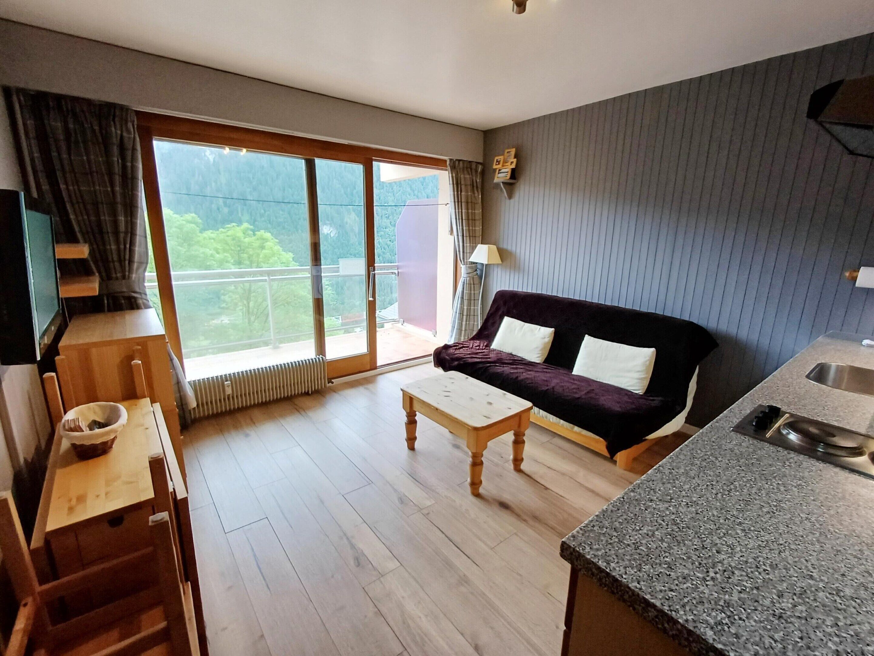Ein modernes Wohnzimmer mit einer Couch, einem Couchtisch, einer Küchenzeile mit Spüle und Blick durch eine große Schiebeglastür.