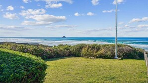 A grassy area with a tall white pole, a wooden fence, and a view of the ocean with waves and a distant ship. - Falcon Getaway - New Oceanfront Listing! (Arch Cape)