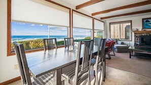 A dining area with a large table and chairs, large windows with blinds, a fireplace, and a view of the ocean. - Falcon Getaway - New Oceanfront Listing! (Arch Cape)