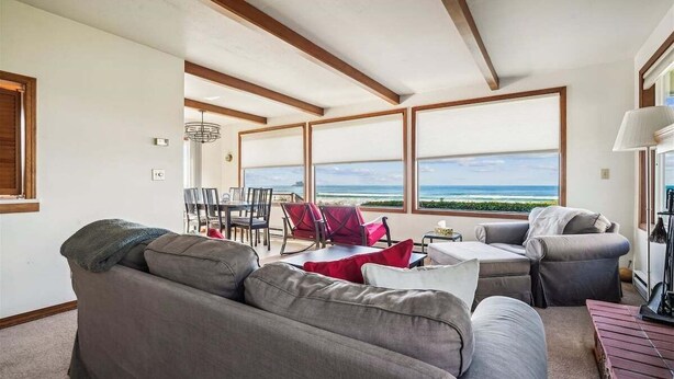 A spacious living room with a large gray sofa, a dining table with chairs, and a view of the ocean through large windows. - Falcon Getaway - New Oceanfront Listing! (Arch Cape)