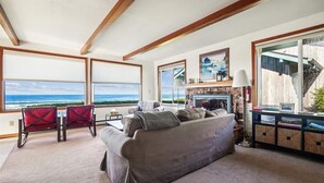 A living room with a large window overlooking the ocean, a fireplace, and a cozy seating area. - Falcon Getaway - New Oceanfront Listing! (Arch Cape)
