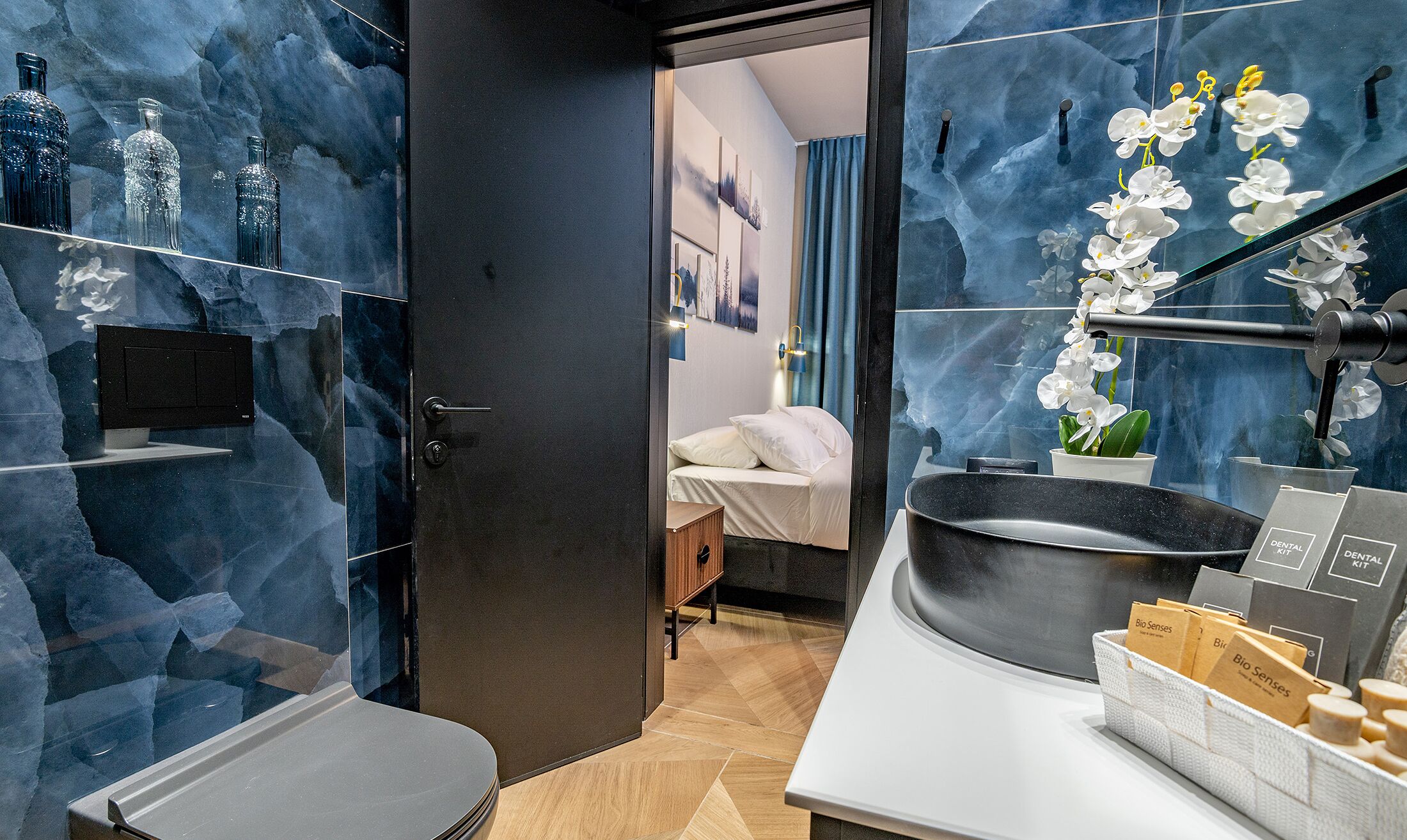 Ein modernes Badezimmer mit einem großen Spiegel, einem Waschbecken und einer Toilette. Regale mit Pflegeprodukten und ein Topf mit einer Orchidee sind ebenfalls vorhanden.