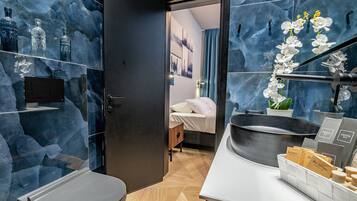 Ein modernes Badezimmer mit einem großen Spiegel, einem Waschbecken und einer Toilette. Regale mit Pflegeprodukten und ein Topf mit einer Orchidee sind ebenfalls vorhanden.