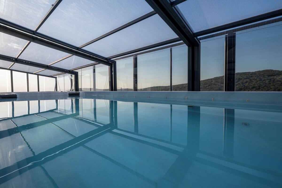 Une grande piscine intérieure aux parois en verre transparent, offrant une vue sur un paysage vallonné.