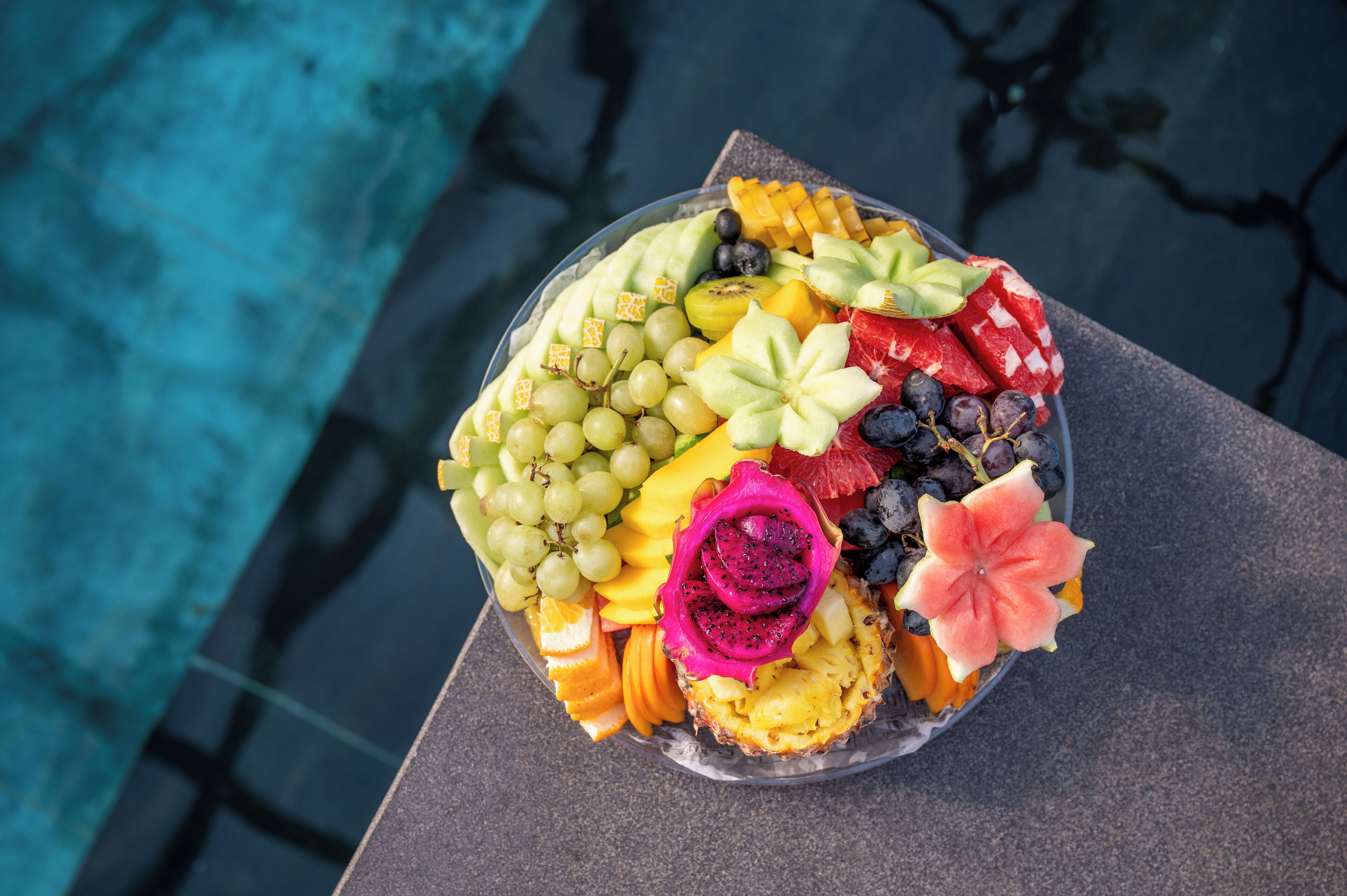 Een schaal met verschillende soorten fruit en een bloemstuk, geplaatst op een tafel nabij een zwembad.