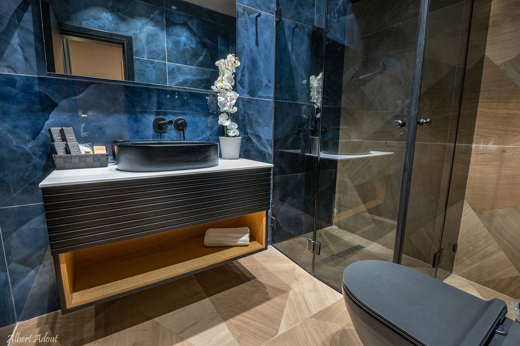 Un baño moderno con un lavabo negro, paredes de mármol, un mueble de tocador de madera y una cabina de ducha de vidrio.