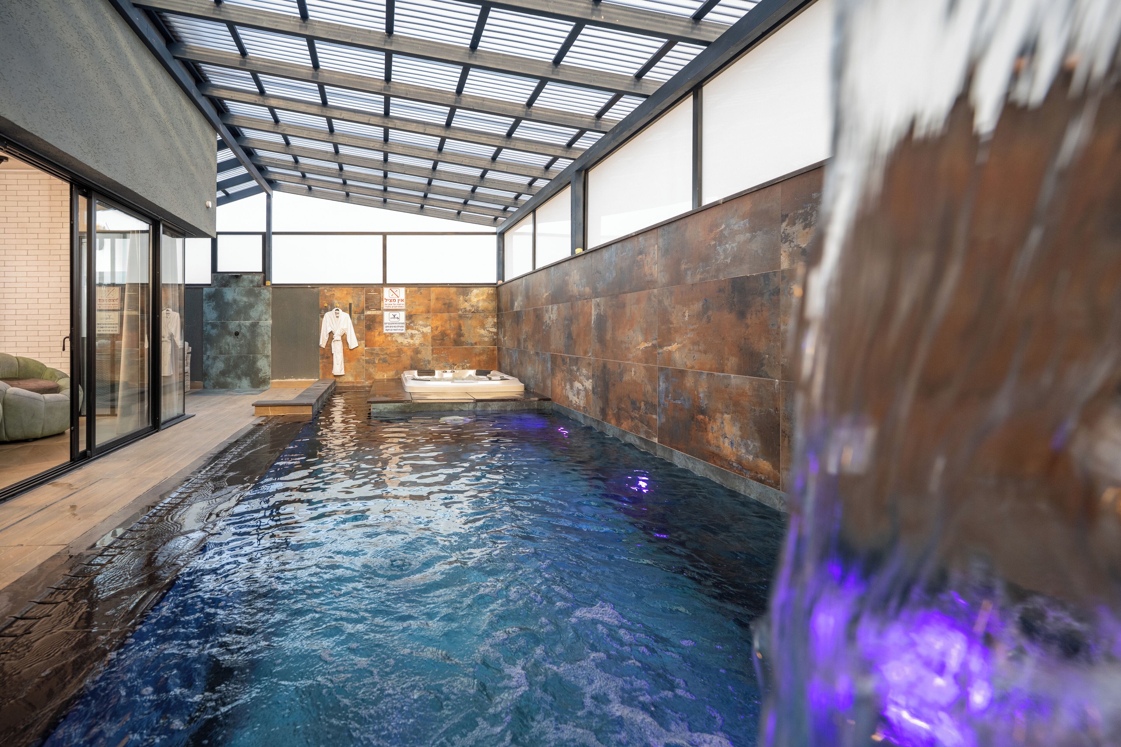An indoor swimming pool with a glass ceiling, a brick wall, and a white robe hanging on the wall.