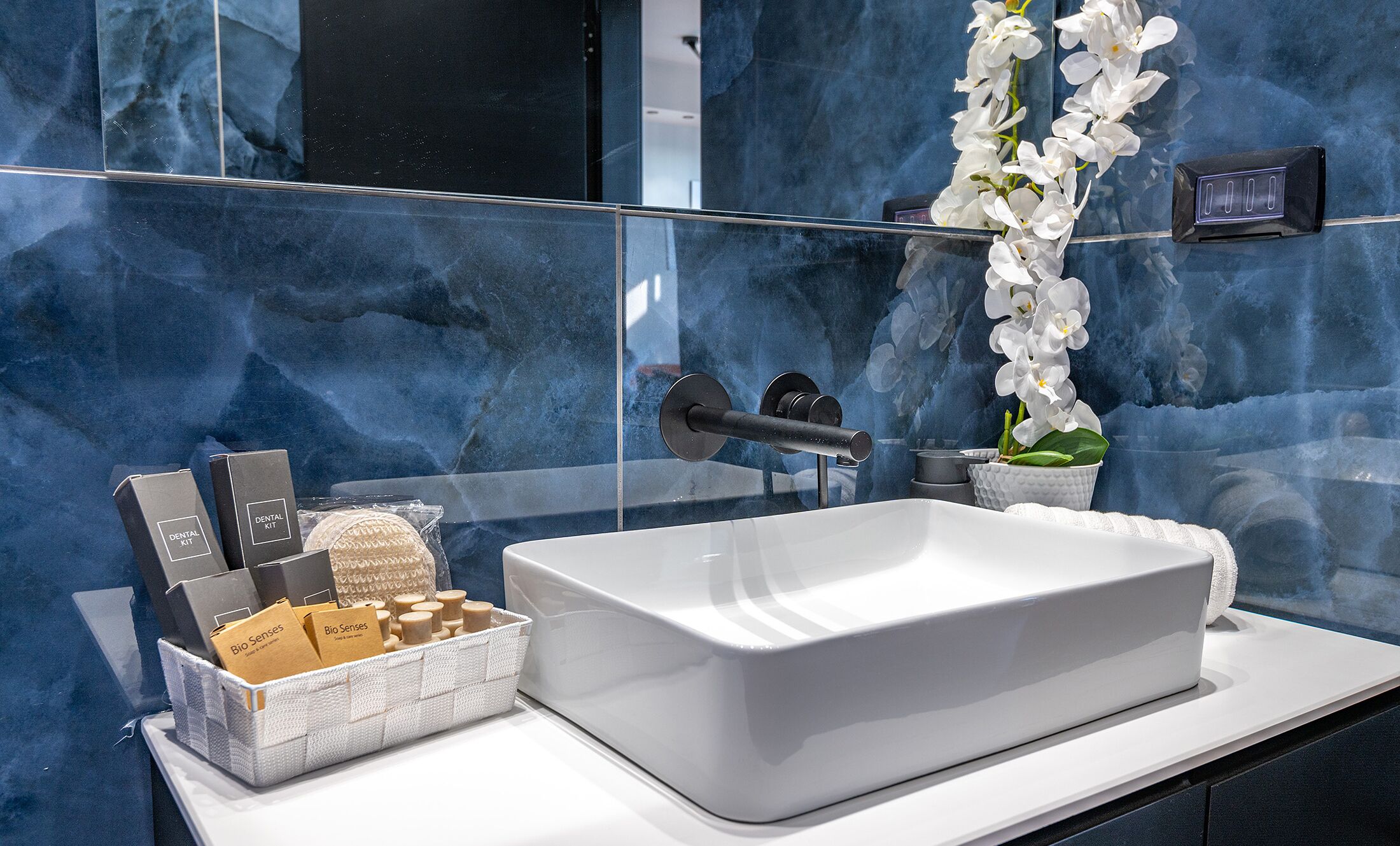 A bathroom with a white sink, a mirror, and a glass partition. There are toiletries in a basket and a white orchid in a vase.