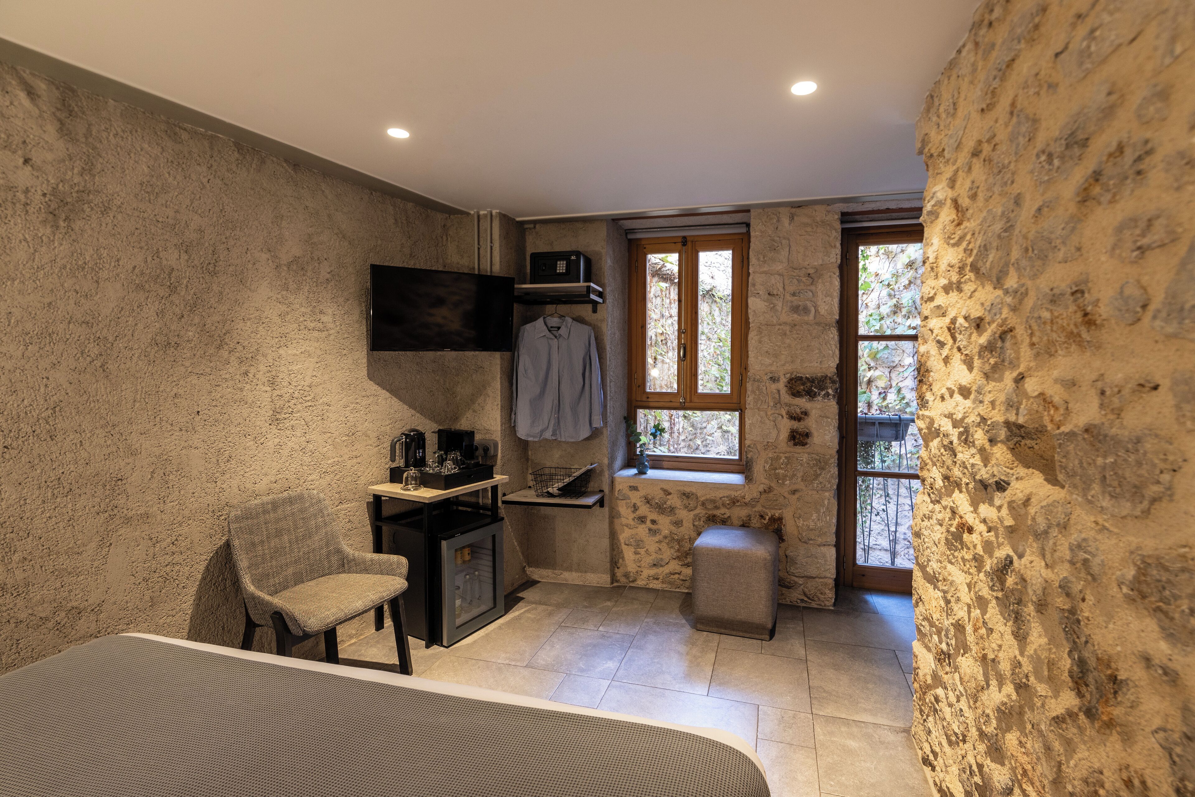Habitación compacta con pared de piedra, una kitchenette pequeña, una silla, una cama y un ventanal que ofrece luz natural.
