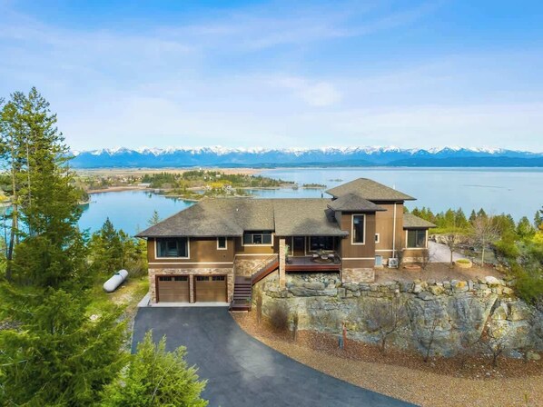 A two-story house with a stone foundation, a garage, and a view of a lake and mountains.