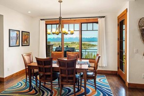 A dining area with a view of mountains and a lake, wooden chairs, a chandelier, and a patterned rug.
