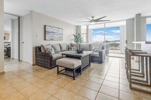 Relax in this spacious living room with a dining area and seating space, featuring a ceiling fan and large windows offering a view of the outdoors.