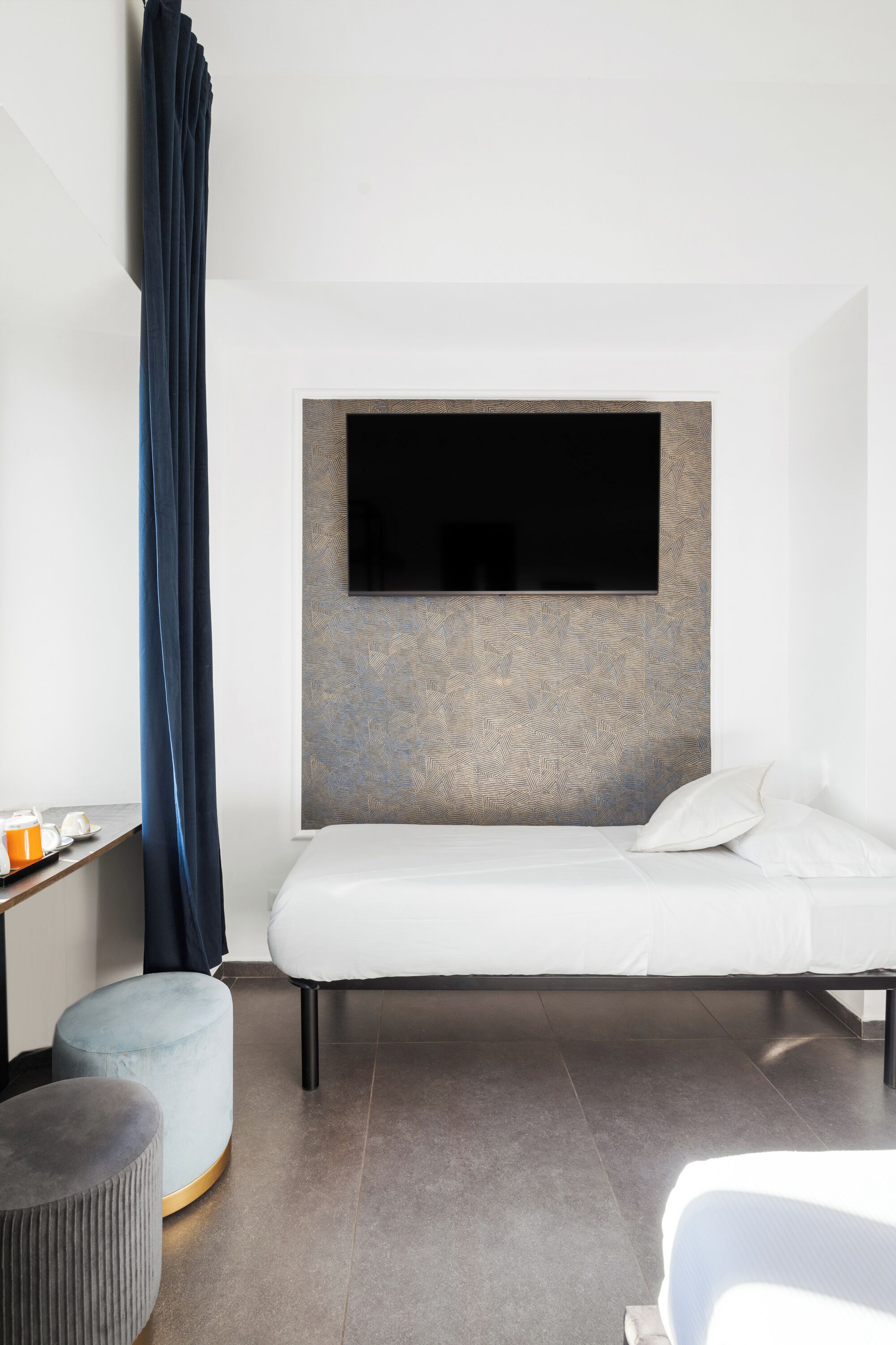 A modern hotel room with a large bed, a mounted TV, a small round ottoman, and a bedside table with a cup and saucer.