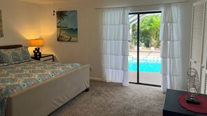 A bedroom with a bed, a ceiling fan, a painting of a beach scene, a window with white curtains, and a view of a swimming pool.
