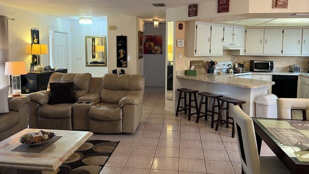 A modern living room with a beige sofa, a dining table with stools, and a kitchen area with white cabinets and a microwave.