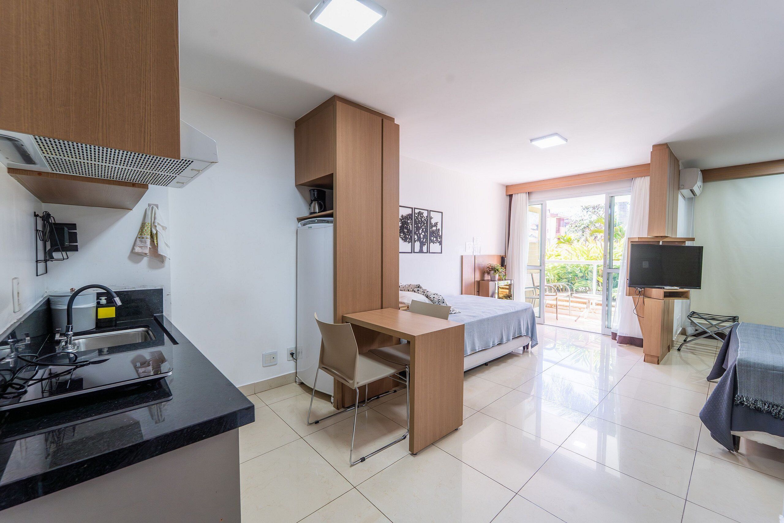 A compact hotel room with a kitchenette, a bed, a TV, and a balcony with a view.