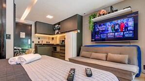 A hotel room with a bed, a sofa, a TV displaying a show, and a kitchenette. - The Sun flat (Brasília)