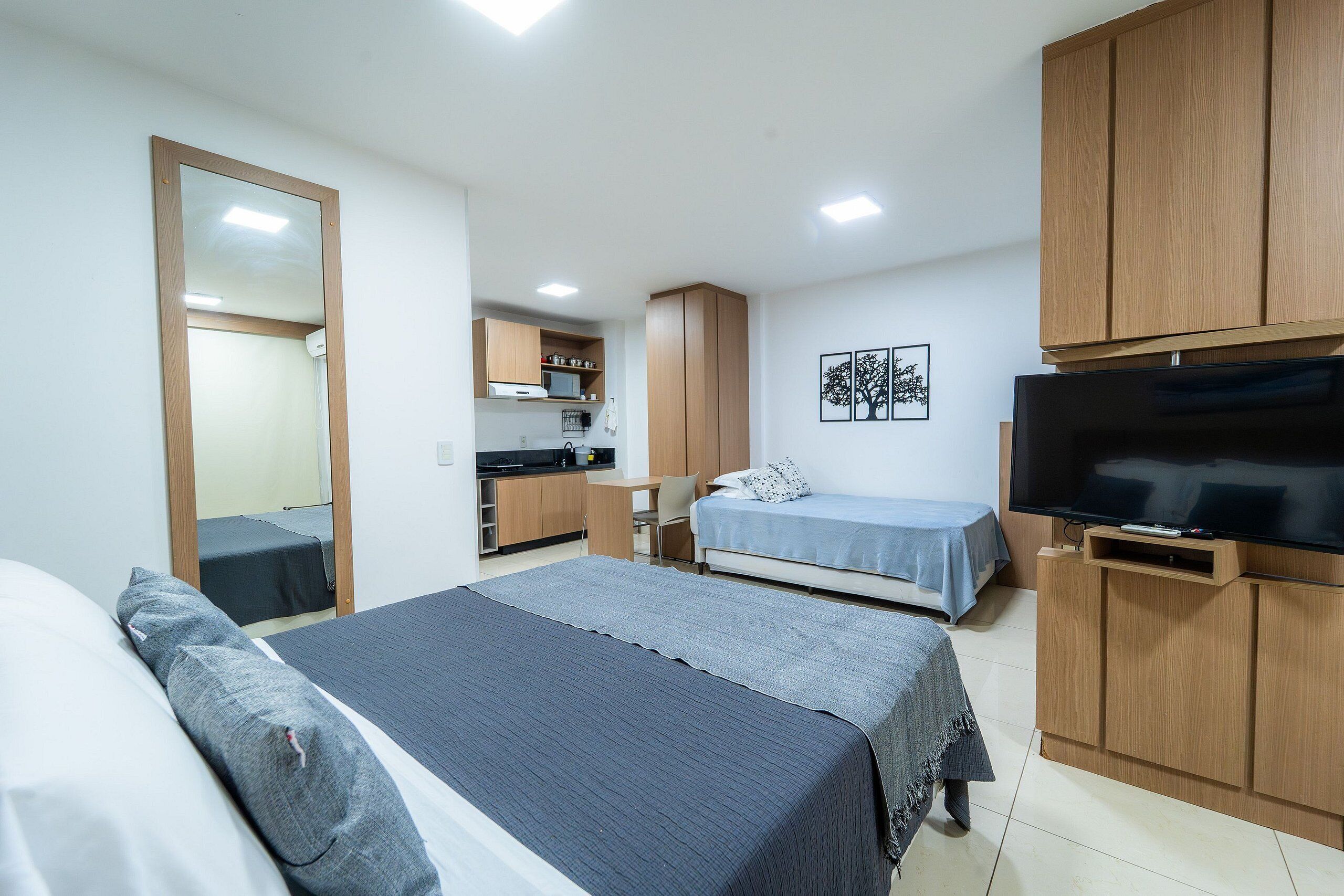 A bedroom with two beds, a TV, wooden cabinets, and a kitchenette in the background.