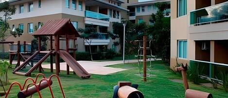 A playground with slides and climbing structures, surrounded by a building with multiple balconies and a flat roof.