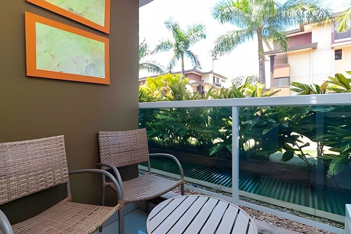 A balcony with wicker furniture, a glass railing, and a view of a swimming pool and buildings.