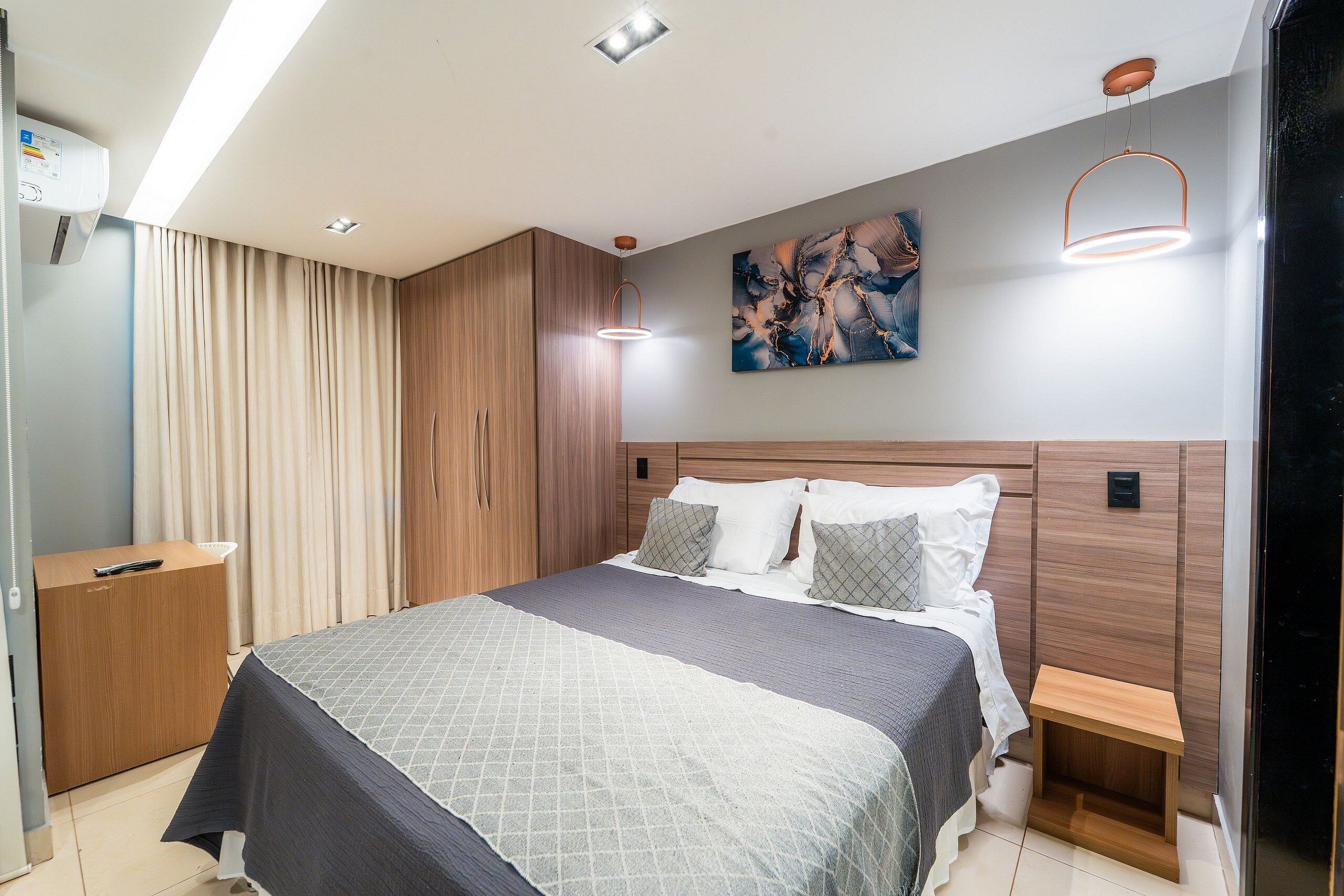 A modern bedroom with a large bed, wooden headboard, a bedside table, a nightstand, a painting on the wall, and a ceiling light fixture.