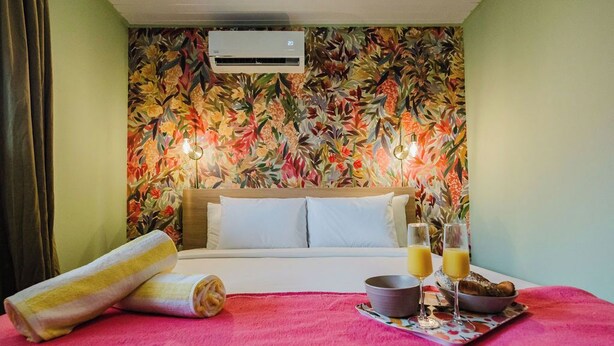 Ein Hotelzimmer mit floralem Wandpapier, einem Bett mit weißer Bettwäsche, einem rosa Teppich, einem Handtuch, einem Tablett mit Frühstücksgegenständen und einer Klimaanlage.
