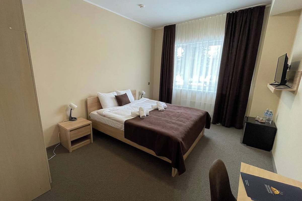 Une chambre d’hôtel comprenant un lit, une table de chevet, un bureau, une chaise, une télévision et une fenêtre avec des rideaux.