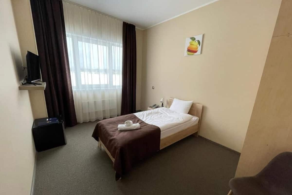 Une chambre d’hôtel comprenant un lit, une chaise, un bureau avec un ordinateur, une fenêtre avec des rideaux et un tableau accroché au mur.