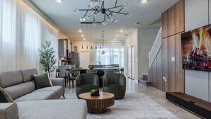 A modern living room with a sofa, armchairs, a coffee table, and a TV. There is a staircase leading to another level.