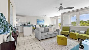 Een moderne woonkamer met een grijze bank, groene fauteuils en een geel poufje. Er is een plafondventilator, een eethoek met een glazen tafel, en een keuken op de achtergrond.