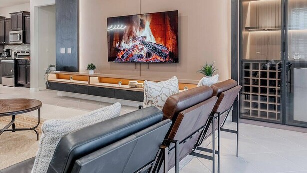 A modern living room with a large flat-screen TV mounted on the wall, a comfortable seating area with leather sofas, a kitchen area with dark cabinets and a microwave, and a glass door leading to a wine cellar.