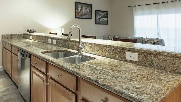 A kitchen with granite countertops, stainless steel sink, wooden cabinets, and a dishwasher. - 4BR house with pool near Disney area (Kissimmee)