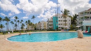 A poolside area with lounge chairs, a swimming pool, palm trees, and a building in the background. - On Bavaro Beach - Secure, Gated Condo Community. In Center of Bavaro-Punta Cana (La Altagracia Province)