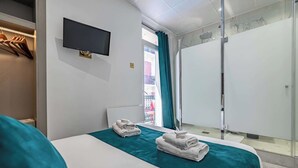 Hotellihuone, jossa on sänky, seinälle asennettu televisio ja kylpyhuone, jossa on lasinen suihkuovi.