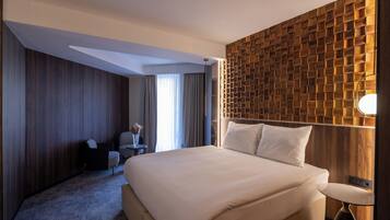 Quarto de hotel moderno com uma cama grande, uma parede de destaque em madeira, uma pequena mesa com abajur e vista para o exterior pela janela.