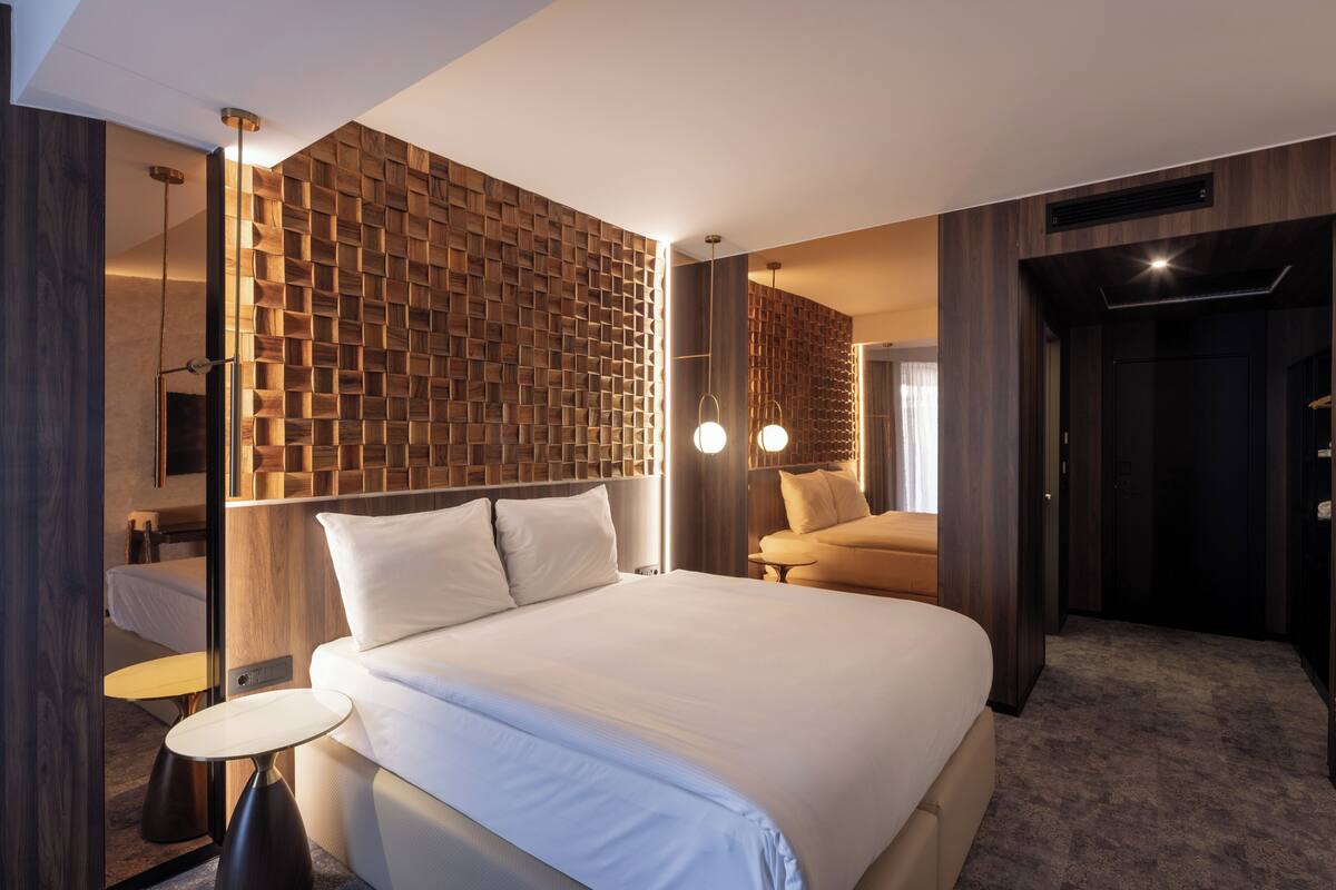 A modern hotel room with a large bed, a round side table, a wooden accent wall, and a separate seating area with a sofa.
