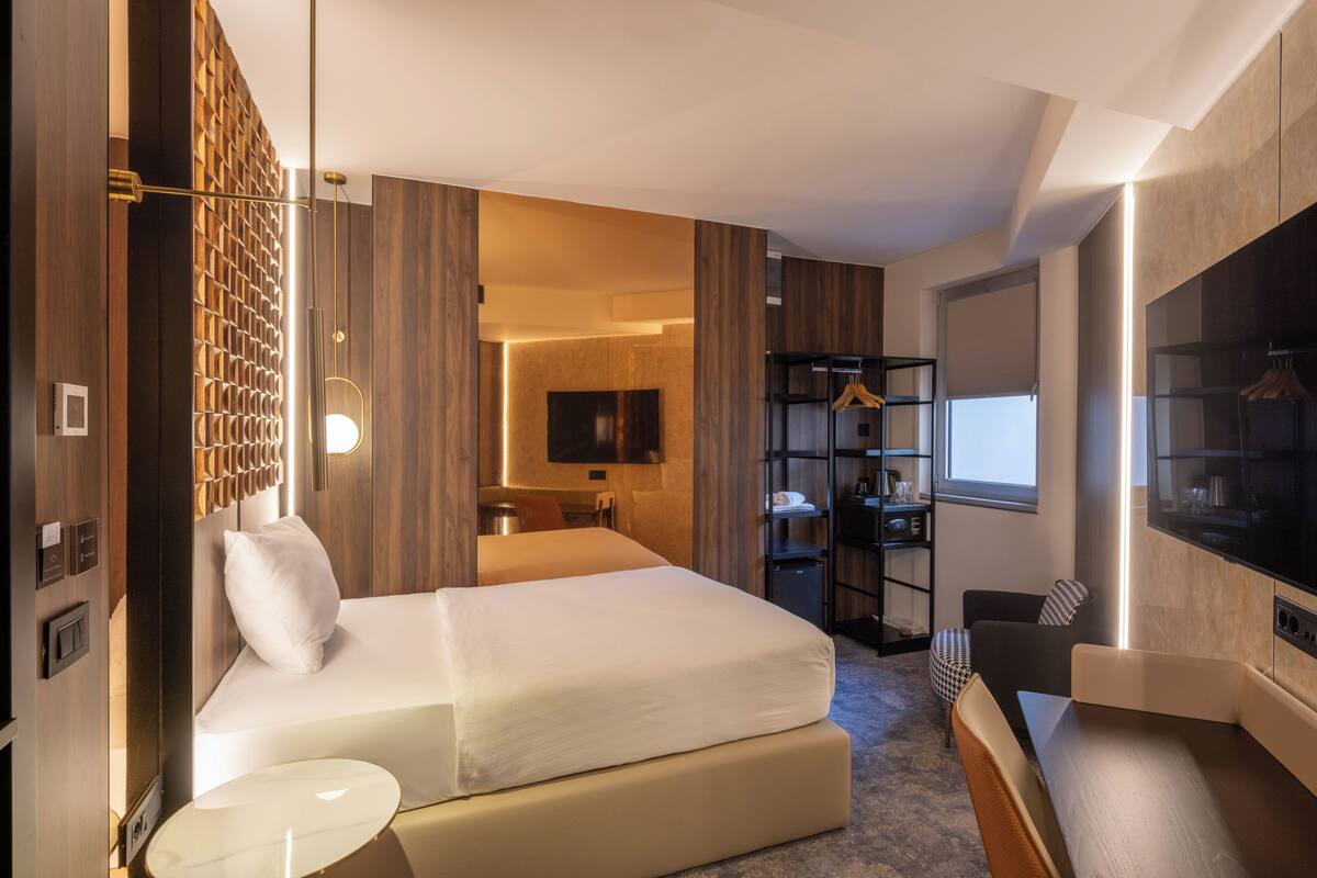 A modern hotel room with a large bed, a TV, a seating area, and a wardrobe.