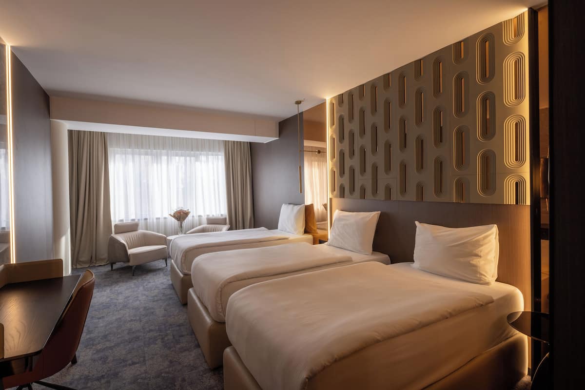 A hotel room with two beds, a desk, a chair, and a large window with curtains.